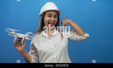 Young hispanic woman engineer wearing a hardhat holding a drone and making a call me hand gesture in studio; confidence innovation. Stock Photo