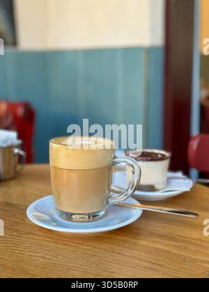 A glass of layered tiramisu is subtly blurred in the foreground, while a clear mug of latte sits on a white saucer on a wooden table, with bright blue Stock Photo