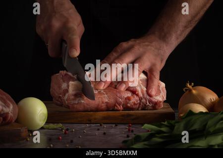 A person prepares fresh meat by slicing it on a wooden board in a dark kitchen. On the table, onions, spices, and bay leaves are arranged, enhancing t Stock Photo