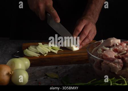 A chef skillfully slices vegetables on a wooden cutting board while raw meat sits in a bowl nearby. Fresh ingredients are laid out, highlighting the c Stock Photo