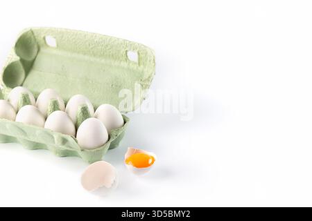 A carton filled with fresh white eggs sits on a clean surface. One egg is broken, revealing a bright yellow yolk against a white backdrop, showcasing Stock Photo