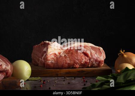 Pork roast rests on a wooden board alongside an onion, green leaves, and scattered spices. The dark background highlights the fresh ingredients, sugge Stock Photo