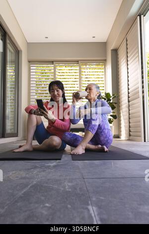 Asian woman and elderly woman relaxing on yoga mats, using smartphone and drinking Stock Photo
