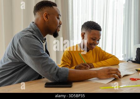 Father helping son with homework at home, both smiling and engaged in learning Stock Photo