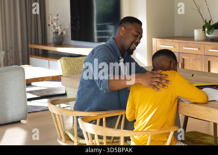 African American father assisting son drawing at wooden table in modern living room using notebook Stock Photo