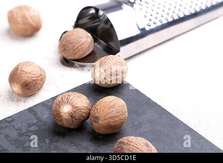 Whole nutmegs, on a stone tile, next to a nutmeg grater, on linen. A few dried seeds of fragrant or true nutmeg fruits, Myristica fragrans. Stock Photo
