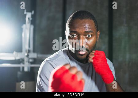 Workout in gym. African man fighter ready for fight punching with boxing wraps protective bandages to camera. Strong man training punches looking concentrated straight preparing for boxing sparring || Model released Stock Photo