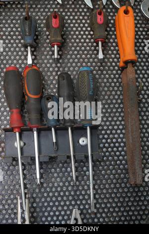 A vertical shot of various hand tools, including screwdrivers and a file with a rusty blade, neatly organized on a dark, perforated workshop pegboard. Stock Photo