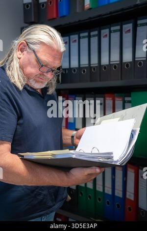 Mature Caucasian man with glasses, next to a filing cabinet in an office reading documents. Stock Photo