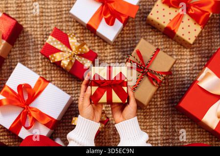 Small present with red ribbon and bow in hands above the gift boxes on the floor. Stock Photo