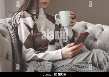 Woman relaxing home on sofa, holding mug and using smartphone while wrapped in warm beige knitted blanket. Cozy winter atmosphere with calm indoors, s Stock Photo
