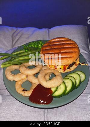 A close-up of a meal featuring a grilled burger with melted cheese, onion rings, peas, cucumbers, and sauce on a plate. Captured indoors in Alicante, Stock Photo