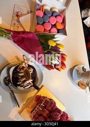 A box of colorful macarons paired with a bouquet of tulips rests on a bright table along with desserts and coffee. Stock Photo