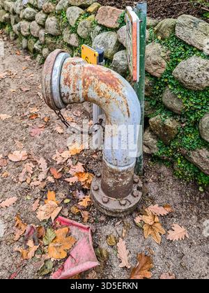 Rusty industrial pipe with a chain and bolts emerging from the ground, surrounded by colorful autumn oak leaves, contrasting with a weathered stone wa Stock Photo