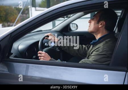 A man sits in the driver s seat, focused on the road with both hands on the steering wheel, captured from the side inside a modern car. Stock Photo