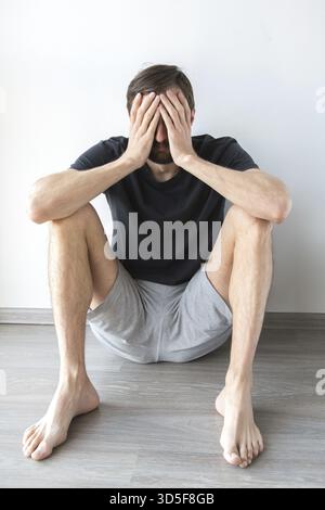 Frustrated young man sitting on the floor with his hands covering his face, expressing stress and emotional distress Stock Photo