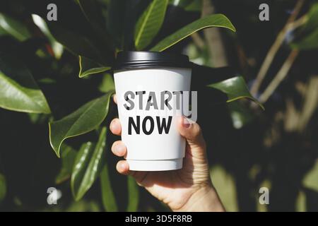 Hand holding a white takeaway cup with bold black text Start Now on background of green tropical leaves in sunlight, concept of motivation Stock Photo