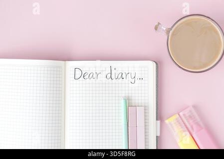 Open notebook with checkered pages and text Dear diary... on pink background, surrounded by pastel pens, highlighters and a cup of coffee with milk, c Stock Photo