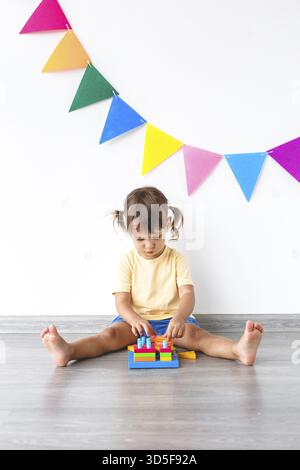 One white girl 2 years old sits on the floor in the room and plays with a multi-colored pyramid sorter on a white background with colored flags, verti Stock Photo