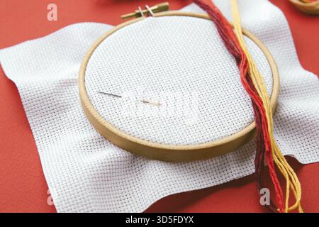 Embroidery hoop with white canvas, two needles and colorful threads on red background, flat lay of cross-stitching tools, concept of handmade craft an Stock Photo