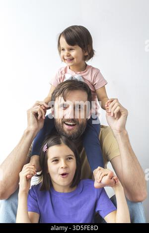 Happy Father with two daughters posing together, smiling having fun. younger girl sits on his shoulders, older one holds his hands on white background Stock Photo