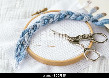 Embroidery tools and materials, wooden hoop, blue embroidery floss, metal scissors, and needles on white fabric, arranged for needlework and crafting Stock Photo