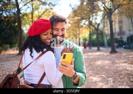 Interracial couple hugging and smiling while looking at smartphone in autumn park Stock Photo