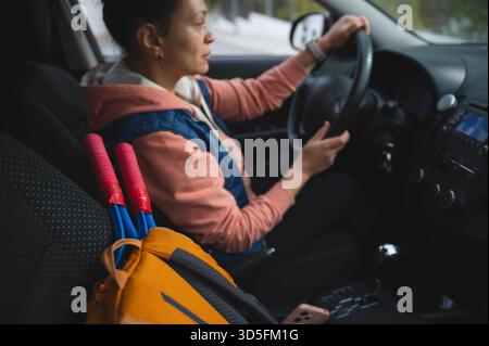 A woman sits behind the wheel of a car, gripping the steering wheel as she drives. A colorful backpack rests in the passenger seat, suggesting travel, Stock Photo