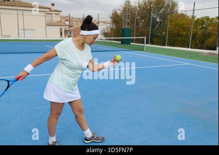 A female tennis player on a blue court prepares to hit a ball with a racket, wearing a light blue top, white skirt, and headband, showcasing focus and Stock Photo