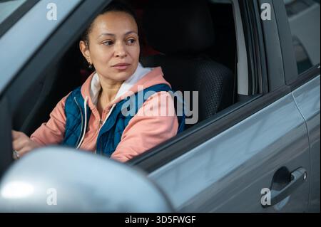 A woman sits in the driver's seat of a car, wearing a pink hoodie and blue vest, looking to the side with a calm, thoughtful expression in a car inter Stock Photo
