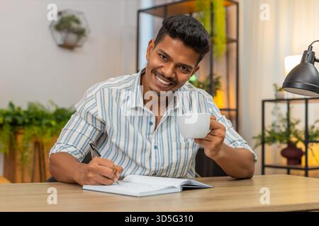 Indian man at home drinks coffee and writes goals in notebook ticks boxes and plans day with drive. Arabian guy at desk reviews list adds tasks sets priorities smiles feels focus and motivation aim Stock Photo