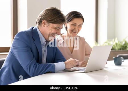 Smiling senior businessman mentor mature businesswoman using notebook at desk Stock Photo