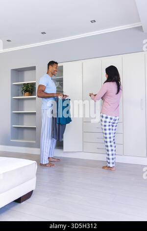 Diverse couple waking up in modern bedroom, holding smartphone and garments by closet storage Stock Photo