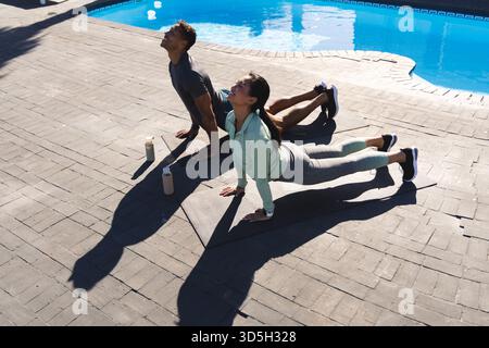 Couple practicing yoga by pool, stretching on mats under morning sun Stock Photo