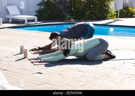 Couple practicing yoga by pool, stretching on mats under morning sun Stock Photo