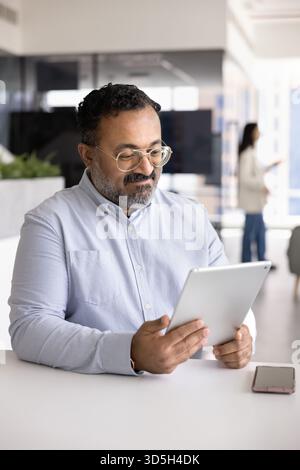 Positive senior Indian businessman reading online content on tablet Stock Photo