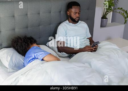 Young couple waking in bedroom, man texting smartphone while woman snoozing on comforter Stock Photo