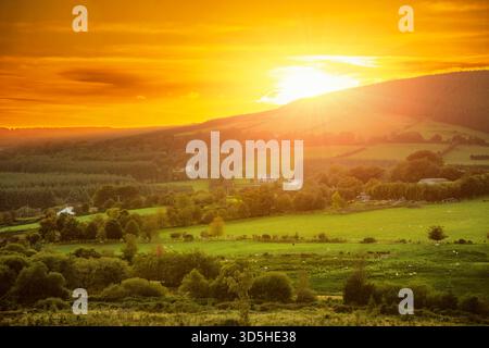 Fiery sunset over Irish valley with green fields, forested hills, and white farmhouses on the autumn equinox Stock Photo
