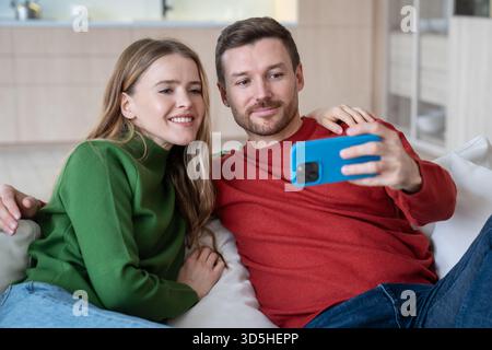 Pleased couple in love take selfies together for social media on cellphone at home capturing moment. Stock Photo