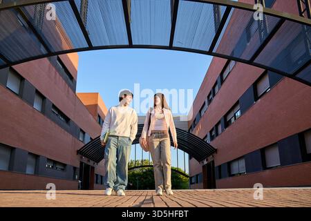 Two young students walking together between modern university buildings, carrying books and backpacks under a clear sky Stock Photo