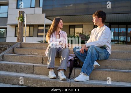 Young male and female students conversing outdoors on concrete stairs, sharing a casual moment between classes on a university campus Stock Photo