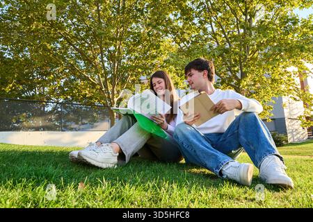 Two young students sitting on green grass, reading and using a tablet, enjoying outdoor learning in a campus park Stock Photo