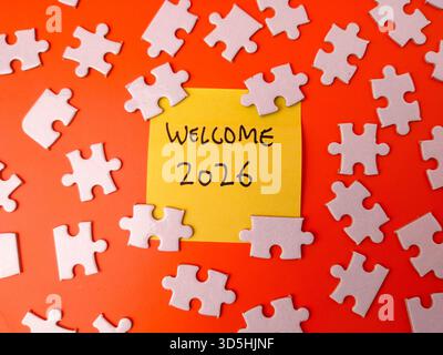 Yellow sticky note with the text WELCOME 2026 surrounded by white jigsaw puzzle pieces on an orange background. Stock Photo