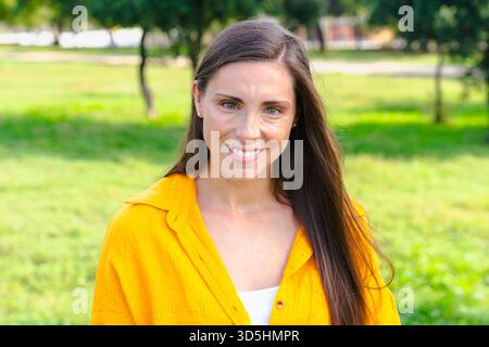 Smiling caucasian woman in yellow shirt looking at camera outdoors in sunny park Stock Photo