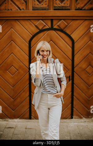 A young woman with blonde hair dressed in a striped shirt and blazer stands outside near a wooden door, smiling as she talks on her phone. Her stylish Stock Photo