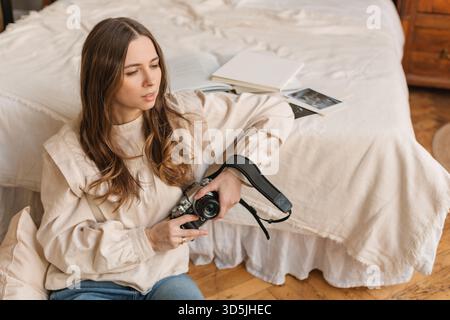 Mid adult woman in jeans sitting on the floor holding a camera and leaning on the edge of her bed Stock Photo