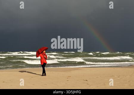 Smiling woman standing on a sandy beach by a rough sea holding a red umbrella on a windy day, Curonian Spit, Lithuania Stock Photo