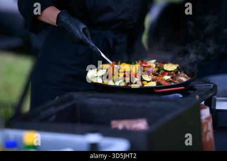 Close-up of a chef standing in a garden wearing black Nitrile gloves cooking assorted fresh vegetables in a frying pan on a gas hob Stock Photo