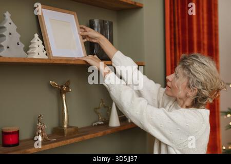 Close-up side view of a Woman arranging an empty picture frame on a shelf amongst assorted festive Christmas decorations Stock Photo