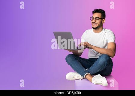 Millennial Arab Man Working on Laptop in Neon Light Setting With Relaxed Posture Stock Photo